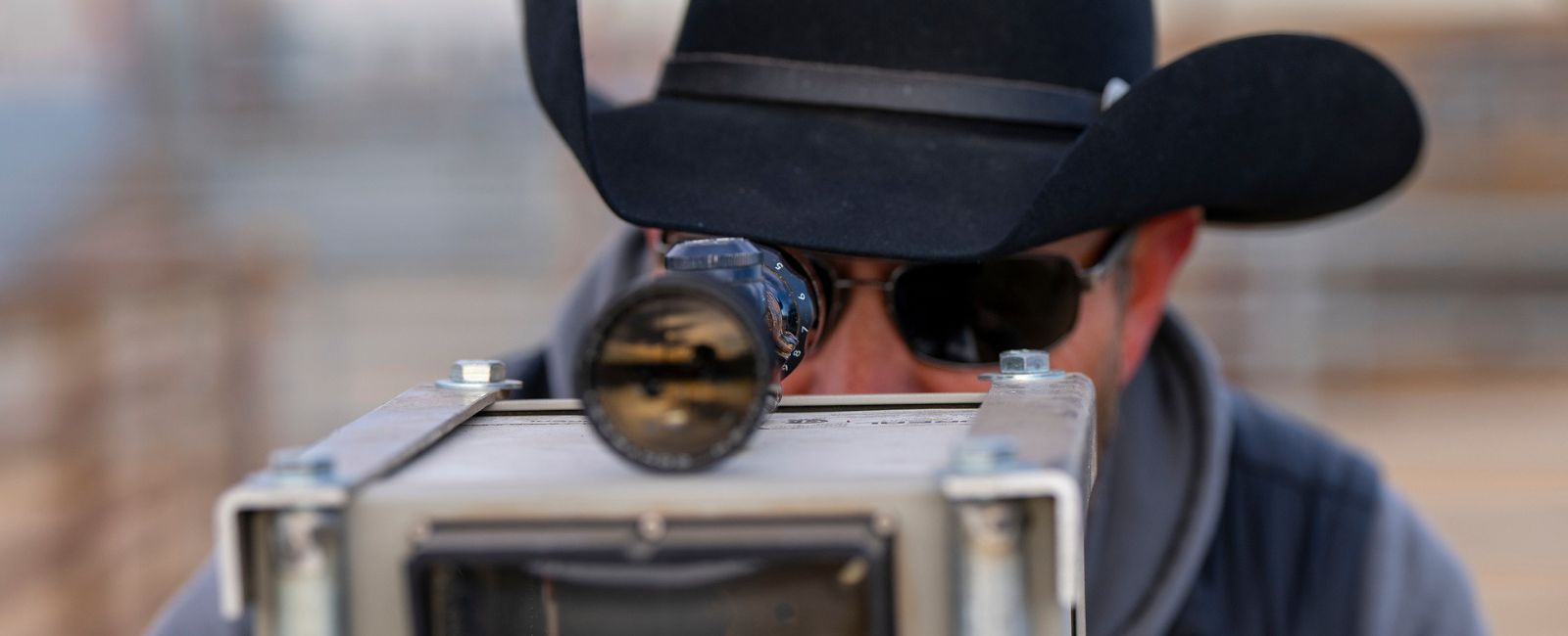 Close-up image of man in cowboy hat looking through a scope attached to a small metal box