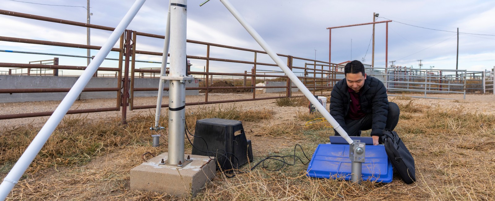 Man kneeling at a laptop computer on  hay-covered ground in a corral in front of a large tripod
